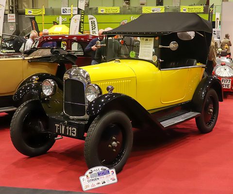 A yellow three-seater with a black hood and plate TW 113 on a red carpet at a busy indoor show. Opens a larger view
