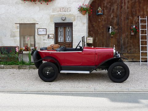 Side view of a red open three-seater with black wings and wheels parked before a stone house with a wooden door. Opens a larger view