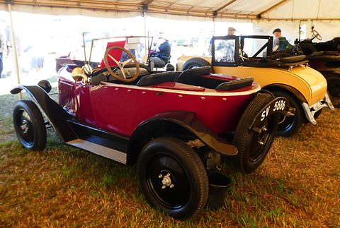 Rear three-quarter view of a red open three-seater with a pointed tail, parked on dry grass under a marquee. Opens a larger view