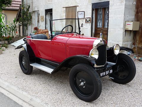 A red open three-seater with black wings and plate 919 SF 39 on gravel before a village house. Opens a larger view