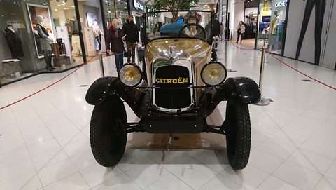 Front view of a tan car with yellow Citroën lettering on the radiator, two lamps and a rope barrier around it. Opens a larger view
