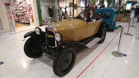 A tan open three-seater with black wings and a Citroën radiator script on a shiny white mall floor. Opens a larger view