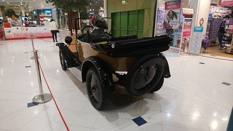 Rear three-quarter view of a tan three-seater with black wings and a folded hood on a white tiled floor. Opens a larger view