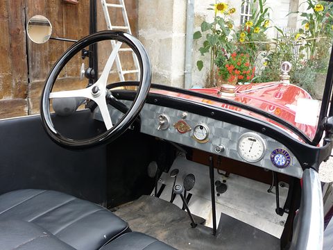 An engine-turned metal dashboard with round gauges, a black steering wheel and pedals on a rubber mat. Opens a larger view