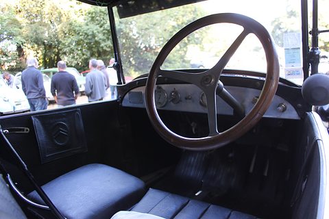 A thin wooden steering wheel before a plain metal dashboard, pedals and a black seat, people outside. Opens a larger view