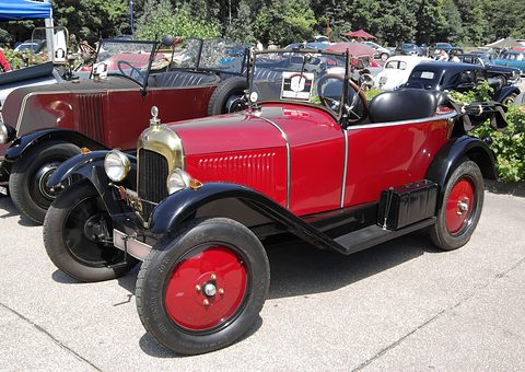 A red open three-seater with a brass radiator and red wheels parked in a row of vintage cars. Opens a larger view