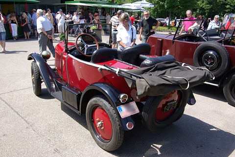 A red open three-seater seen from behind, its folded black hood over the tail and a crowd beyond. Opens a larger view