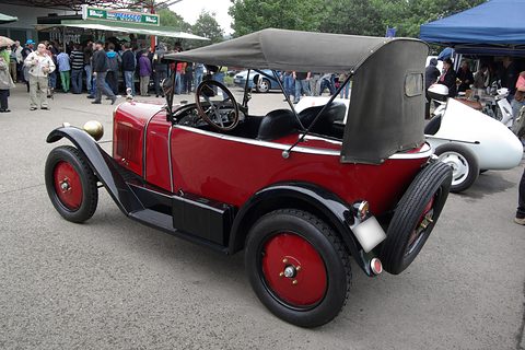 Rear three-quarter view of a red three-seater with a grey hood and a spare wheel on its tail, visitors behind. Opens a larger view