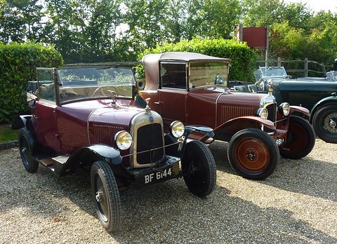 A maroon open cabriolet and a brown one with its hood up, parked together on gravel before a hedge. Opens a larger view