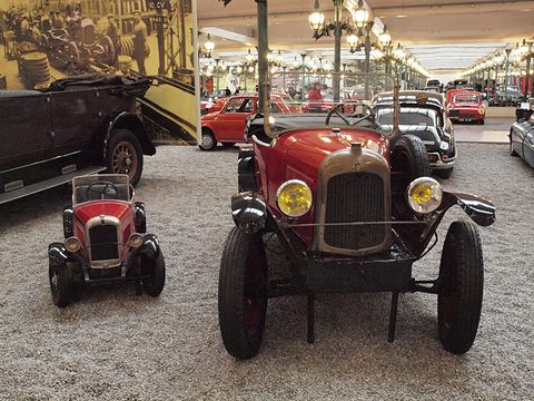 Front view of a red tourer with yellow lamps next to a small red child-size copy on a gravel floor. Opens a larger view