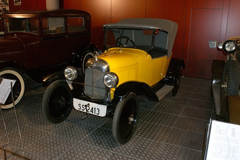 A yellow two-seater with a grey hood and plate SS 2413 on a dark tiled floor beside a Ford. Opens a larger view