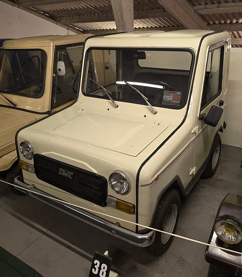 Cream boxy two-seat microcar with a black grille panel, round headlamps and a cab-like body, behind a display rope. Opens a larger view