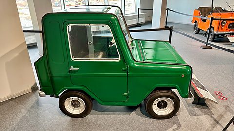 Side view of a green two-seat microcar with a single door, a short bonnet and white wheels, an orange open car beyond.