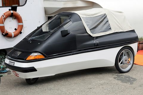 Black and white three-wheeler with a folded cream soft top and alloy wheel beside a camper van with a lifebuoy. Opens a larger view