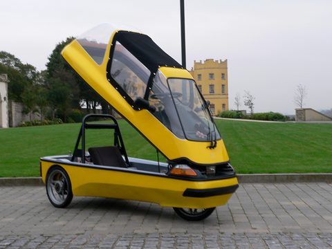 Yellow three-wheeler with its whole glazed upper body tipped forward, parked on paving by a lawn and a tower. Opens a larger view