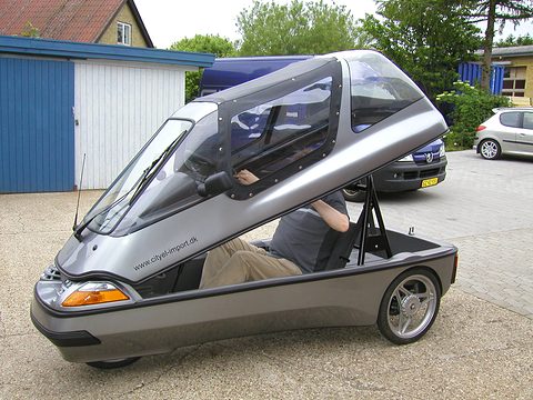 Silver three-wheeler with its glazed upper body hinged up at the nose; a seated man is half hidden inside.