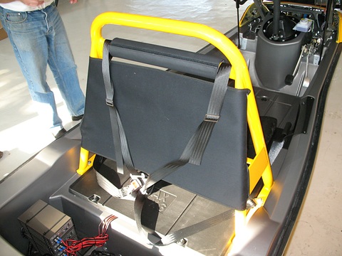 Grey padded child seat with a harness strapped to a yellow roll bar inside an open three-wheeler body.