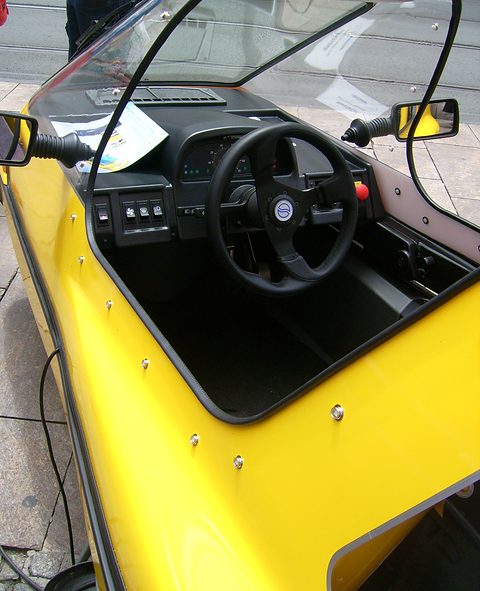 Black three-spoke steering wheel, switch panel and single seat inside a yellow tub, with a door mirror on the frame. Opens a larger view