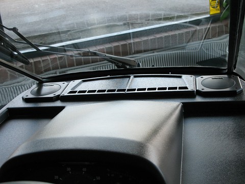Black textured dashboard top with two round speakers and a vent strip under a wet windscreen and wipers. Opens a larger view
