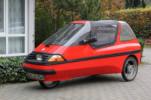 Red three-wheeler with a black grille, amber indicators and soft-top centre section parked in front of ivy and a hedge. Opens a larger view