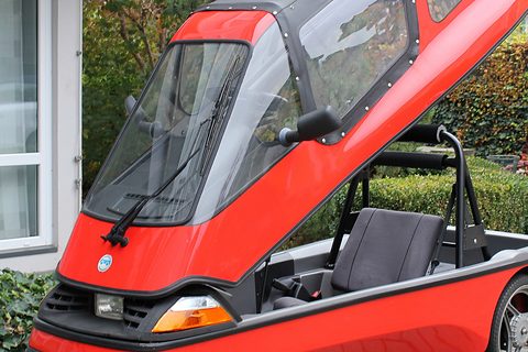 Red glazed upper body tipped up at the nose of a three-wheeler, revealing a black seat and tubular roll bar. Opens a larger view