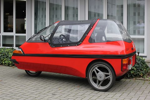 Red three-wheeler with a black fabric centre top and alloy wheel on grey paving in front of large windows. Opens a larger view