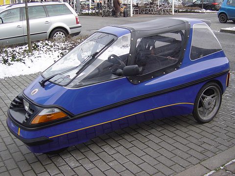 Blue three-wheeler with a black fabric centre top and gold pinstripe parked on paving beside snow-covered shrubs. Opens a larger view