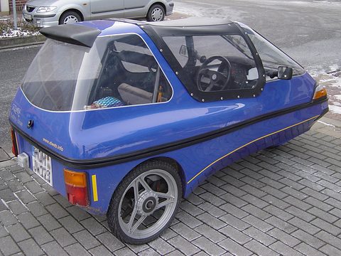 Rear three-quarter of a blue three-wheeler with a black roof spoiler, alloy wheel and number plate on wet paving. Opens a larger view