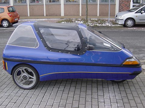 Side view of a blue three-wheeler with a black fabric centre top, alloy rear wheel and glazed rear section. Opens a larger view