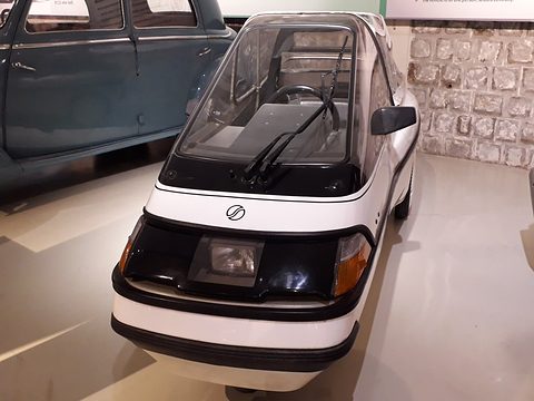 Front of a white single-seat three-wheeler with a glazed canopy, black bumper and central headlamp on a museum floor. Opens a larger view