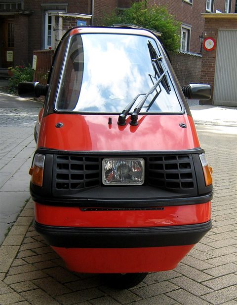 Front of a narrow red three-wheeler with black grille, central headlamp and twin wipers on a brick-paved street. Opens a larger view