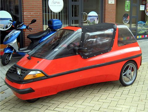 Red three-wheeler with a black fabric top and alloy wheel parked on brick paving beside a blue scooter and shopfront. Opens a larger view