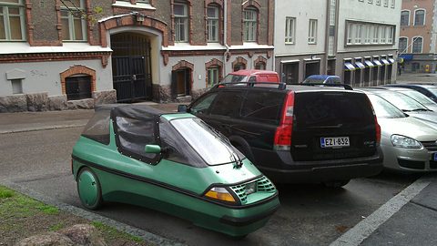 Green three-wheeler with a black soft-top centre section parked at the kerb beside a dark estate car and brick buildings. Opens a larger view