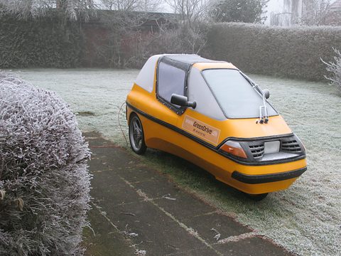 Orange three-wheeler with a white roof and black lower band standing on frost-covered grass beside a white frosted bush. Opens a larger view
