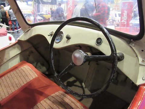 Looking into a cream cockpit with a red and cream tweed bench seat and a black steering wheel. Opens a larger view
