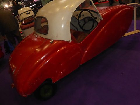 Rear quarter of a red three-wheeler with a cream top, its long rear wing sweeping to the tail.