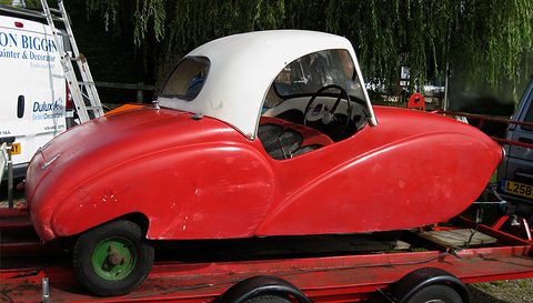 Red streamlined three-wheeler with a white hard top and green wheels on a car trailer under a willow.
