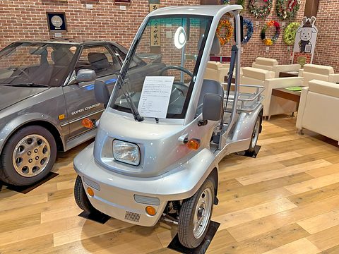 Silver single-seat open microcar with a round headlamp and a tubular rear load rack on a wooden floor.