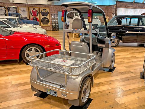 Silver single-seat microcar with a large mesh rear load rack and red tail lamps, classic cars behind. Opens a larger view