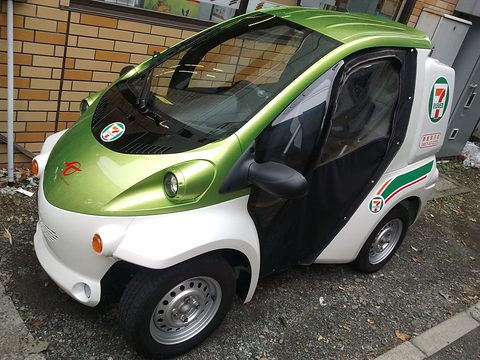Green and white single-seat electric microcar with open sides and a 7-Eleven logo, parked on a gravel lot. Opens a larger view