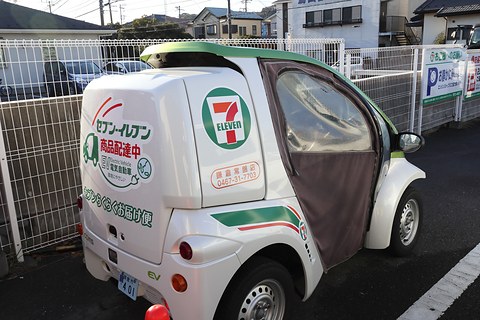 Rear of a white single-seater with a large box lettered in Japanese and a 7-Eleven logo by a mesh fence. Opens a larger view