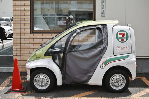Side view of a white and green single-seater with a fabric door curtain and 7-Eleven box, a traffic cone beside it. Opens a larger view