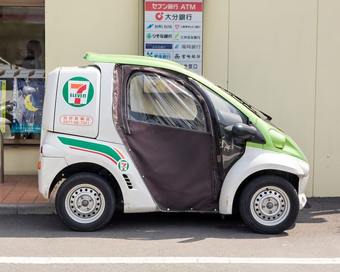Side view of a white and green single-seater with a brown fabric door and 7-Eleven logo by a shop wall.