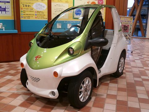 Green and white single-seat microcar with an open side on a terracotta-tiled floor in front of a display board. Opens a larger view