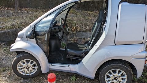 Side view of a silver microcar with no door, showing a black seat, steering wheel and the rear box.