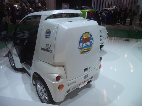 White single-seat microcar with a large rear delivery box bearing a poutine logo, on a glossy white stand. Opens a larger view
