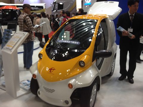 Yellow and white single-seat microcar with its box lid open on a show stand, visitors and a staff member around it. Opens a larger view