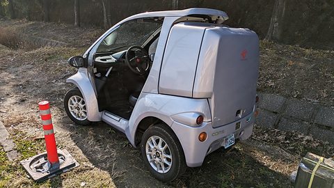 Silver single-seat microcar with a tall rear delivery box beside a red bollard on dry grass. Opens a larger view
