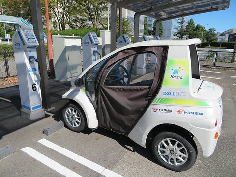 White single-seat microcar with a fabric door and sponsor stickers parked by a row of charging posts. Opens a larger view