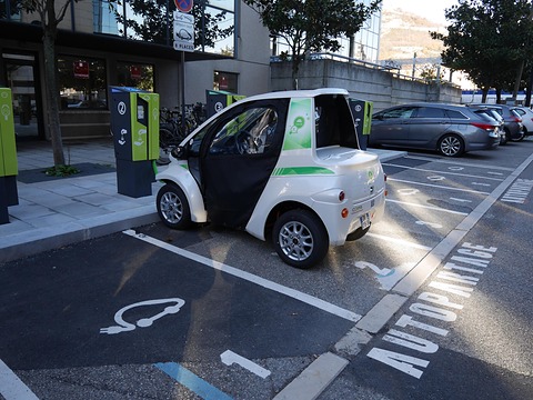 White and green single-seat microcar with an open side parked in a marked charging bay beside charging posts. Opens a larger view